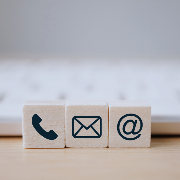Symbolische Holzklötze mit Telefon, E-Mail und @-Symbol vor einer Tastatur.